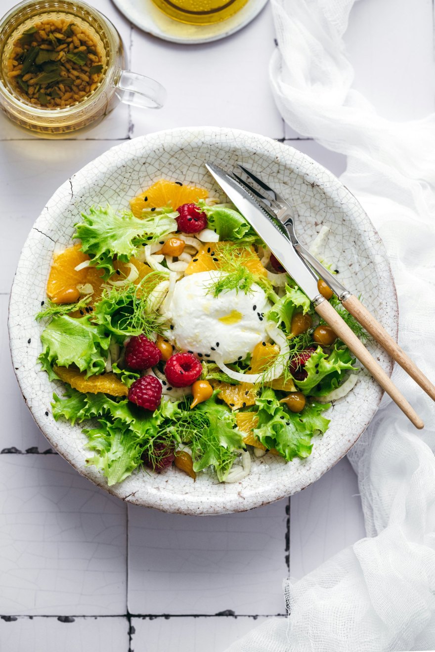 A salad of frisee, orange, raspberries, fennel and burrata in a white ceramic bowl