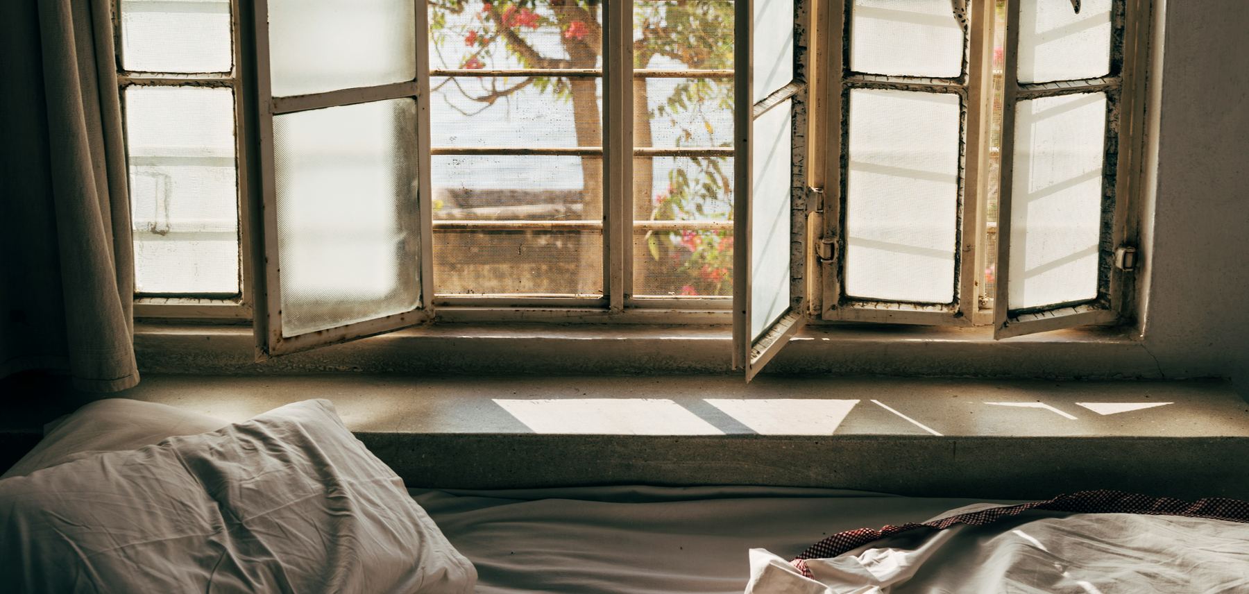 An unmade bed beneath tall windows in soft morning light