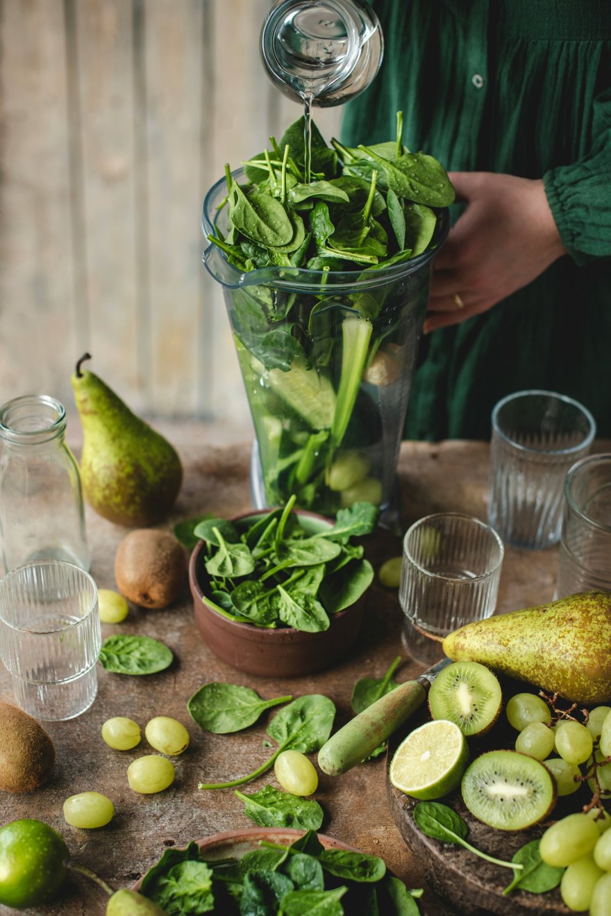 Water being poured into a blender filled with fresh spinach, with pears, kiwi and green grapes on the table