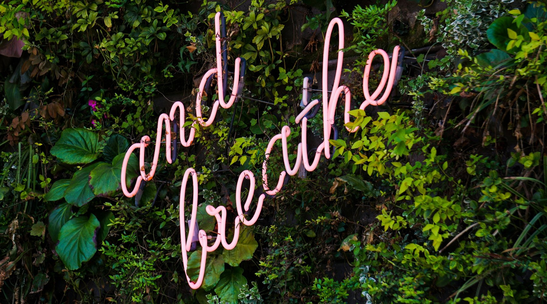 A neon sign reading 'and breathe' on a wall of green plants